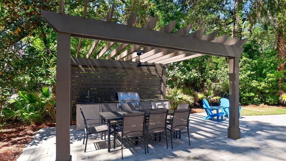 Outdoor dining pergola with a built‑in grill and cozy seating, surrounded by lush greenery and bright blue Adirondack chairs.