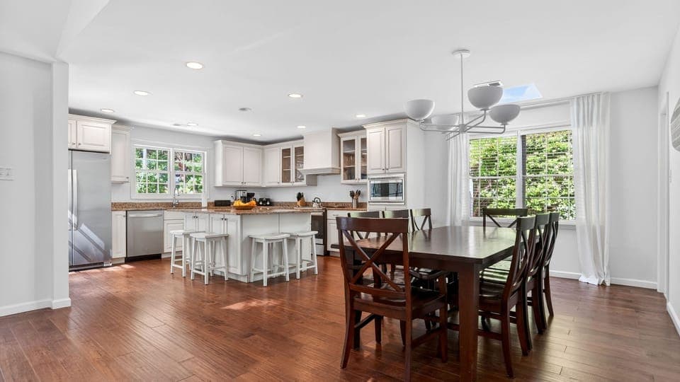 Bright, modern kitchen and dining space with white cabinetry, granite counters, and sun‑filled windows overlooking greenery.