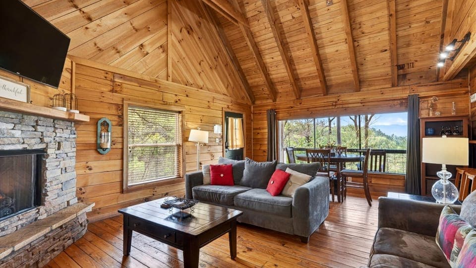 Warm, wood‑lined cabin living room with a stone fireplace, a queen sleeper sofa, soft natural light, and mountain views framed by the dining area windows. (Isaiah's Cabin)