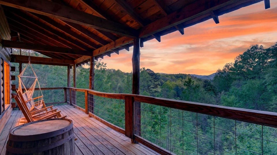 Expansive covered deck with rocking chairs, a hanging swing, and sweeping Smoky Mountain sunset views. (Anna's Cabin)
