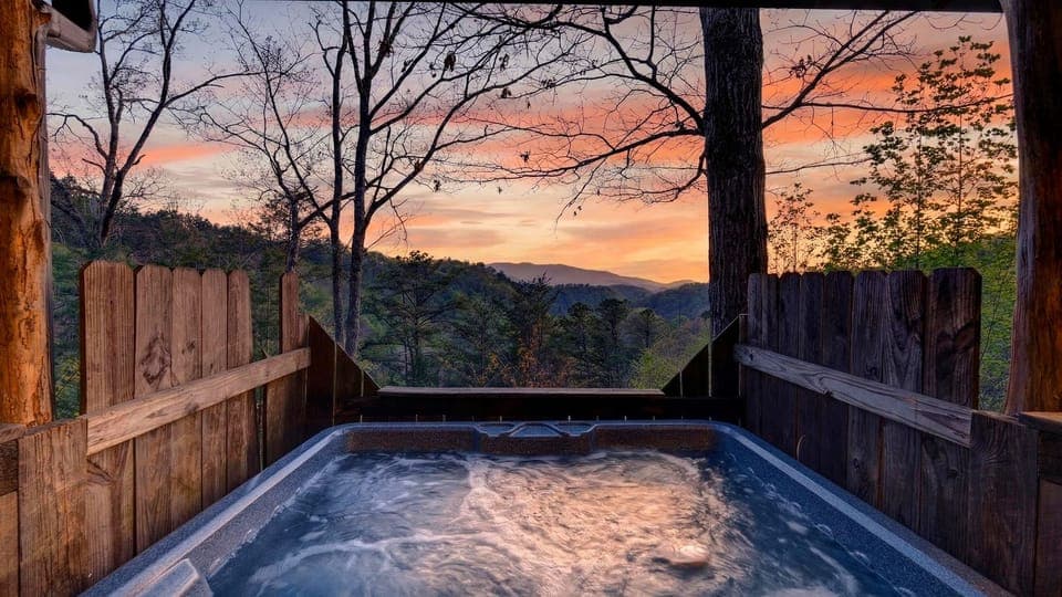 Bubbling hot tub overlooking rolling forested hills under a vibrant sunset sky. (Isaiah's Cabin)