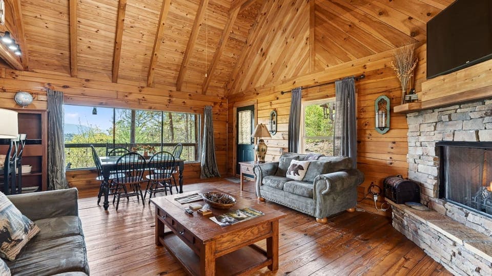 Warm cabin living room with a stone fireplace, cozy seating, and big windows opening to peaceful mountain views. (Jack's Cabin)