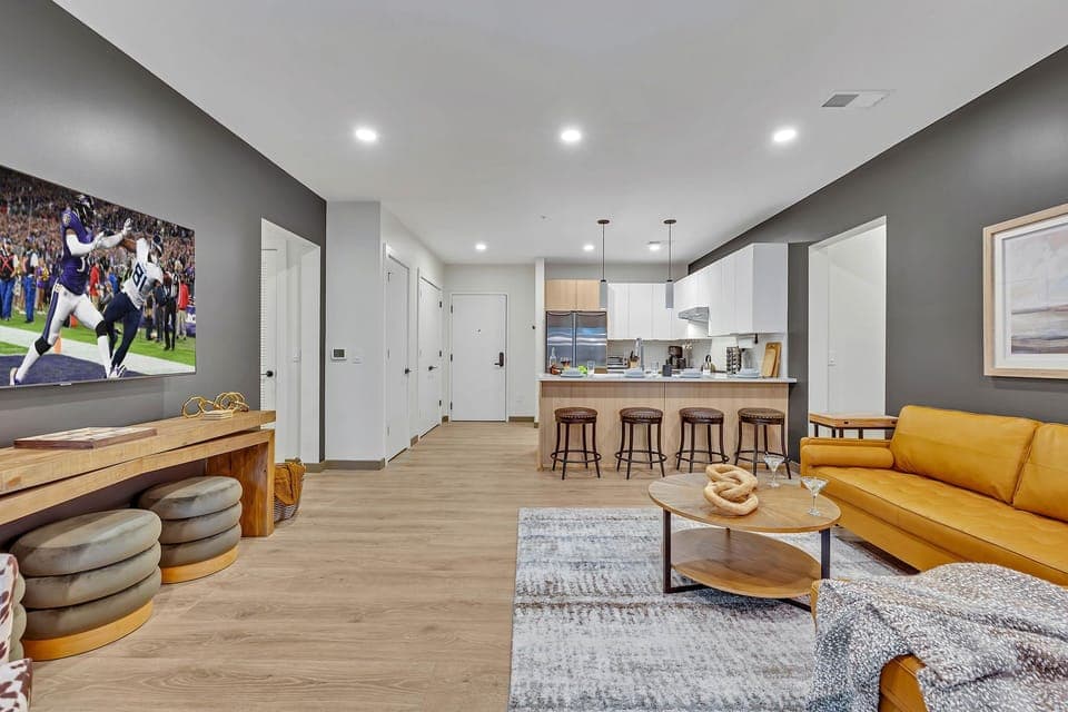 Modern living room decorated in calming tones, featuring comfortable sectional and flat-screen smart TV, and is connected to the kitchen.
