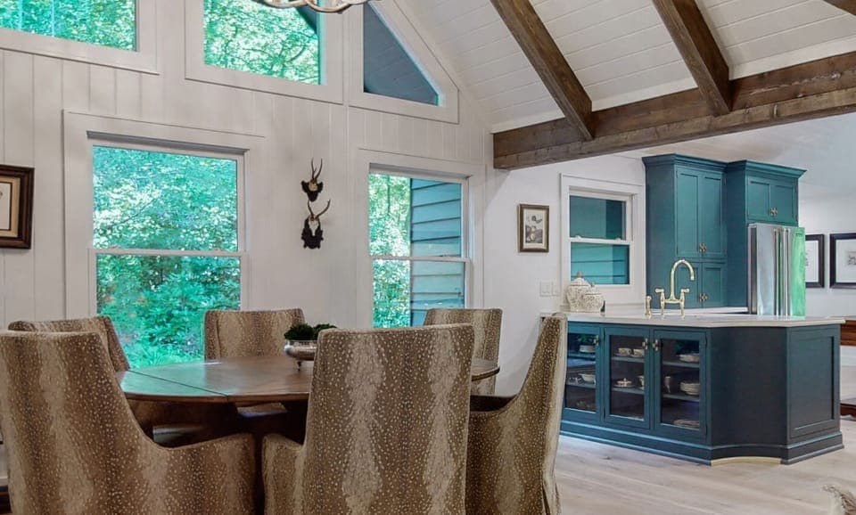 Dining area with large windows and a view of the surrounding greenery next to a kitchen with blue cabinetry and exposed wooden beams.