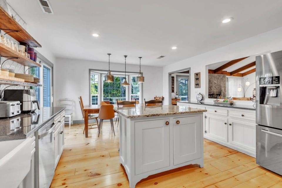 Bright and spacious kitchen featuring modern stainless steel appliances, a granite island, farmhouse-style dining area, and large windows with serene forest views. Fully stocked with cooking essentials, including spices, pots & pans, utensils& more