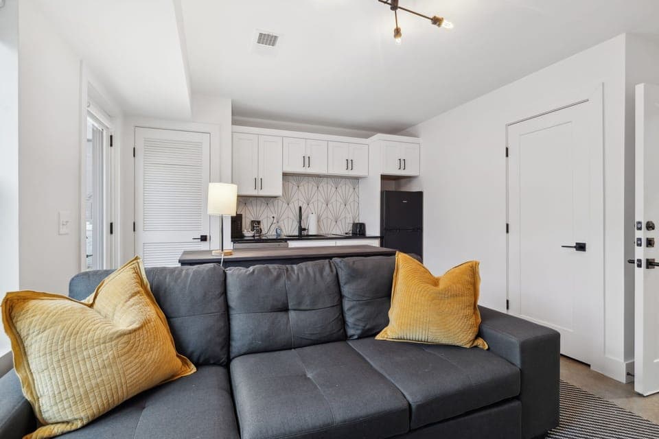 Living Room: Seamless blend of living and kitchen spaces with a cozy sectional, mustard accents, and a fully equipped kitchen.