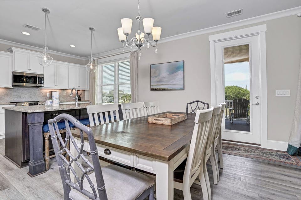 Your elegant dining space features a rustic wooden table beneath a stunning chandelier, with kitchen island seating and balcony doors flooding the room with natural light.