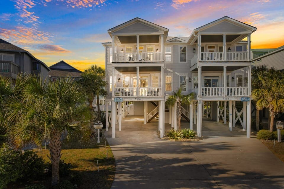 Coastal vacation home with multiple covered balconies and tropical palm trees, photographed during golden hour twilight.