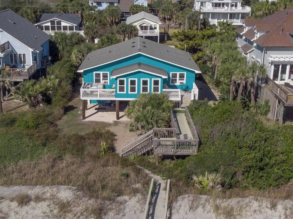 Charming turquoise beach house surrounded by tropical palms, with a wooden boardwalk over coastal dunes.