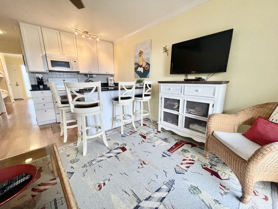 Cozy living area with a kitchen counter, stools, a wall-mounted TV, wicker chair, and a lighthouse-themed rug. White cabinets and a painting decorate the space.