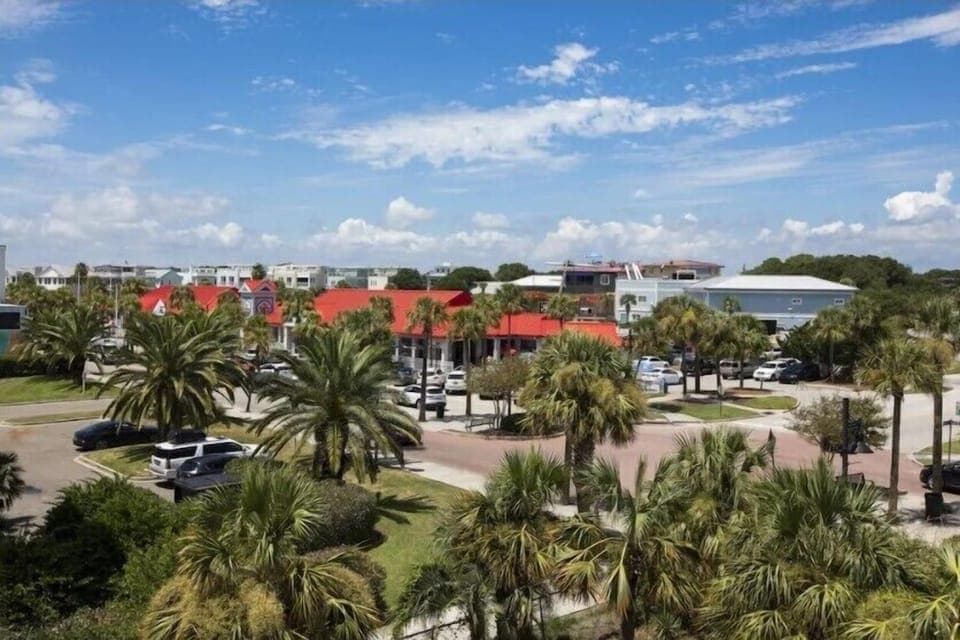 View of a parking lot with palm trees and buildings in the background under a blue sky with clouds.