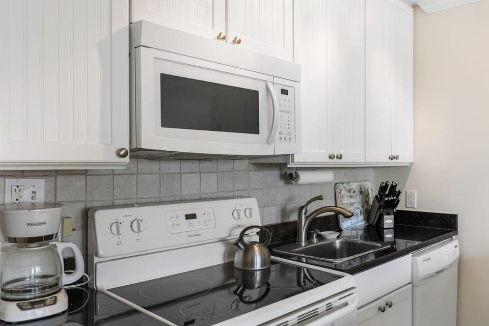 A kitchen with white cabinets, a microwave, a stove, a coffee maker, and a kettle on the countertop. A sink is below the microwave.