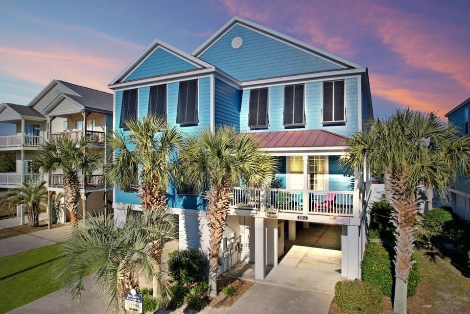 Charming coastal rental with vibrant blue siding, surrounded by swaying palm trees under a stunning sunset sky.