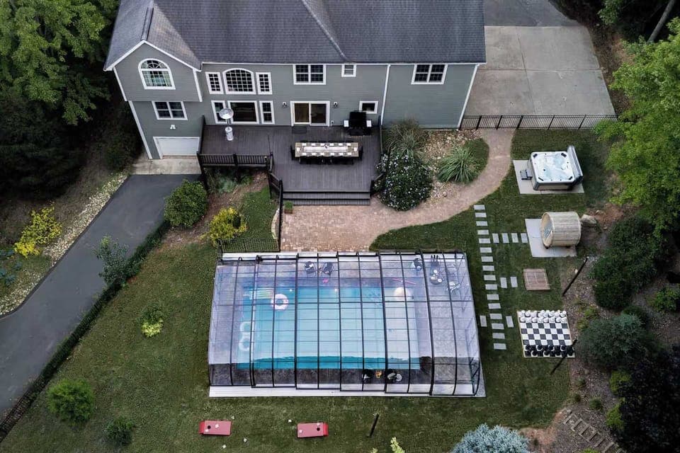 An aerial view of the backyard paradise featuring a heated enclosed pool, hot tub, barrel sauna, life-sized chess set, and cornhole setup.