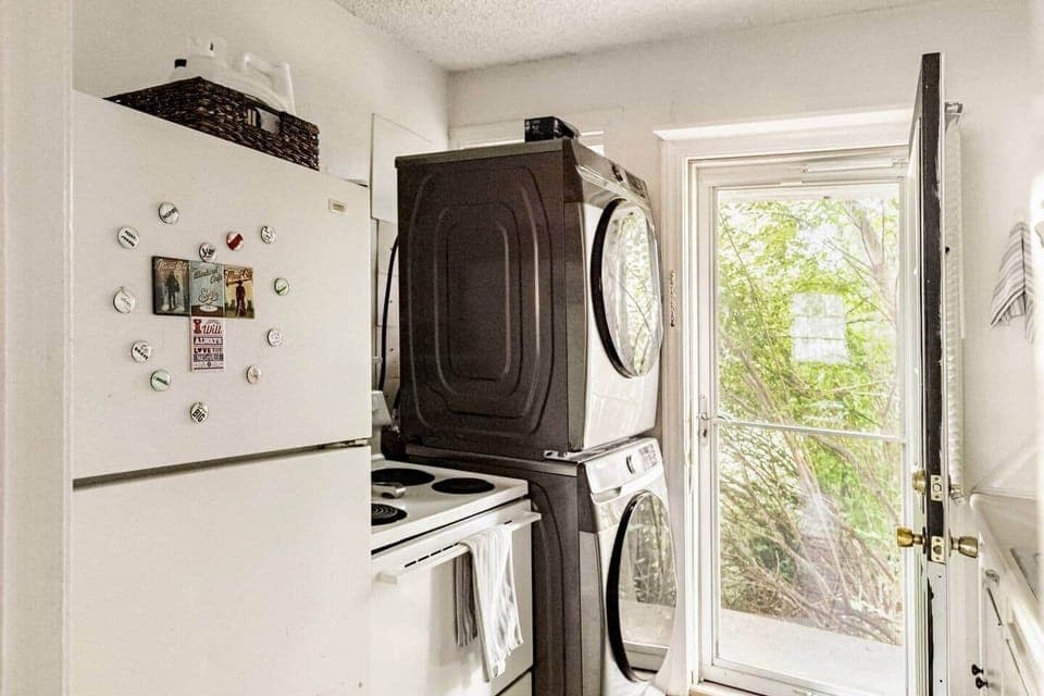 A convenient laundry area with a stacked washer and dryer, a refrigerator with fun magnets, and complimentary laundry detergent & softener. The windowed door brings in natural light, making this space bright and functional for your stay.