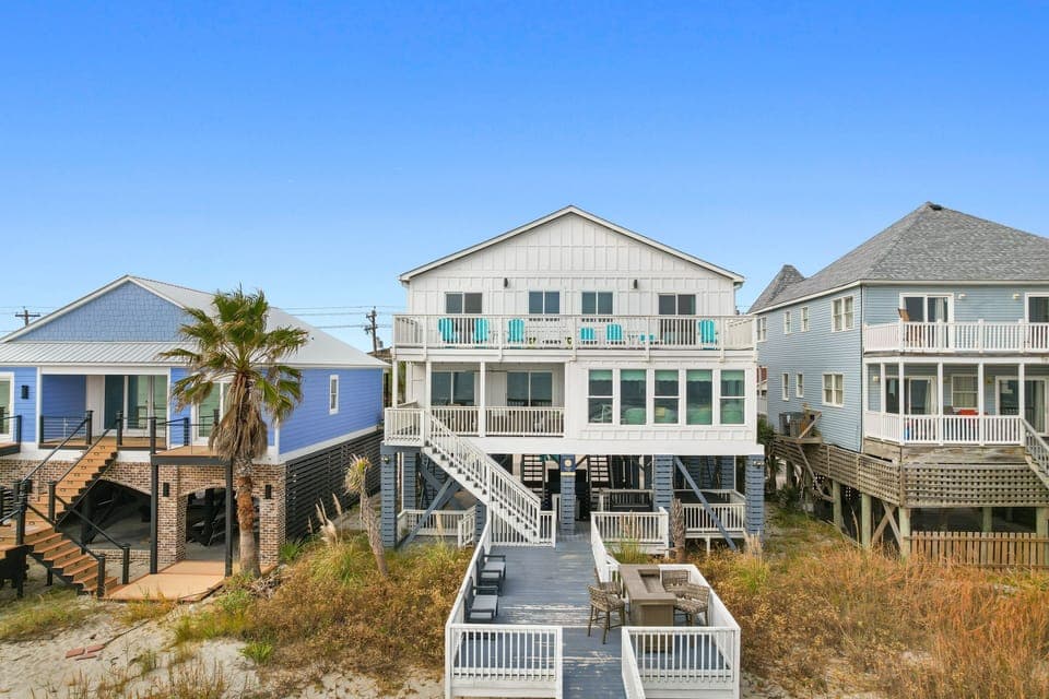 Charming beachfront homes on stilts showcase coastal architecture with multiple decks and balconies against clear blue skies.