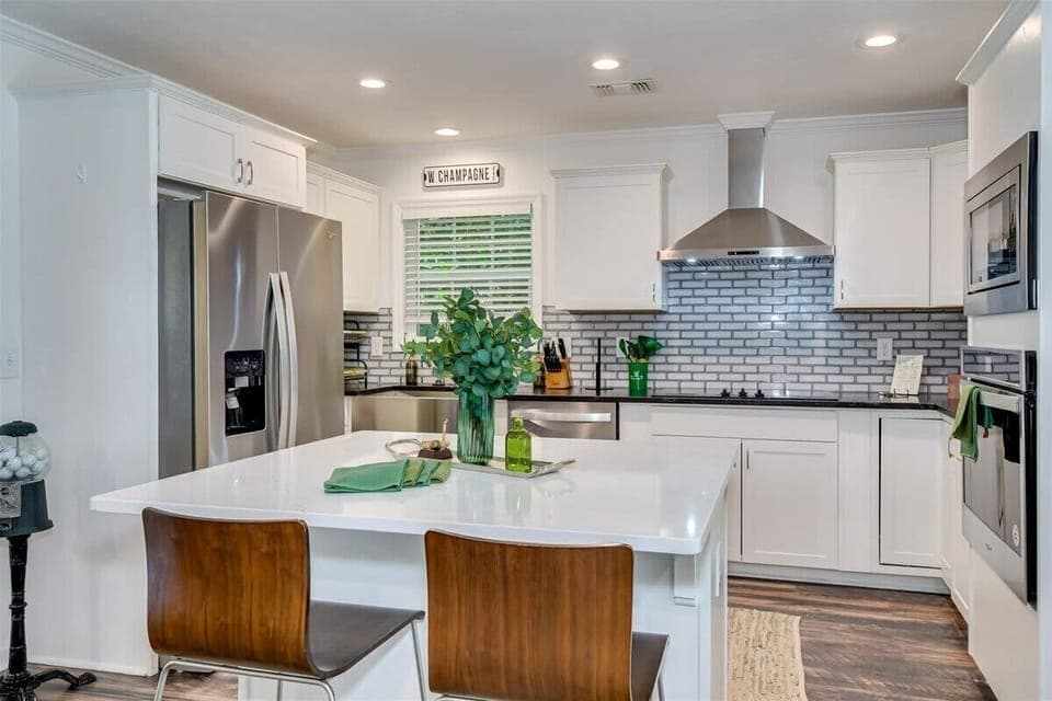 Modern kitchen featuring sleek stainless steel appliances, elegant white cabinetry, and a spacious island with seating. Beautiful backsplash and greenery add a fresh touch, making it perfect for culinary adventures!
