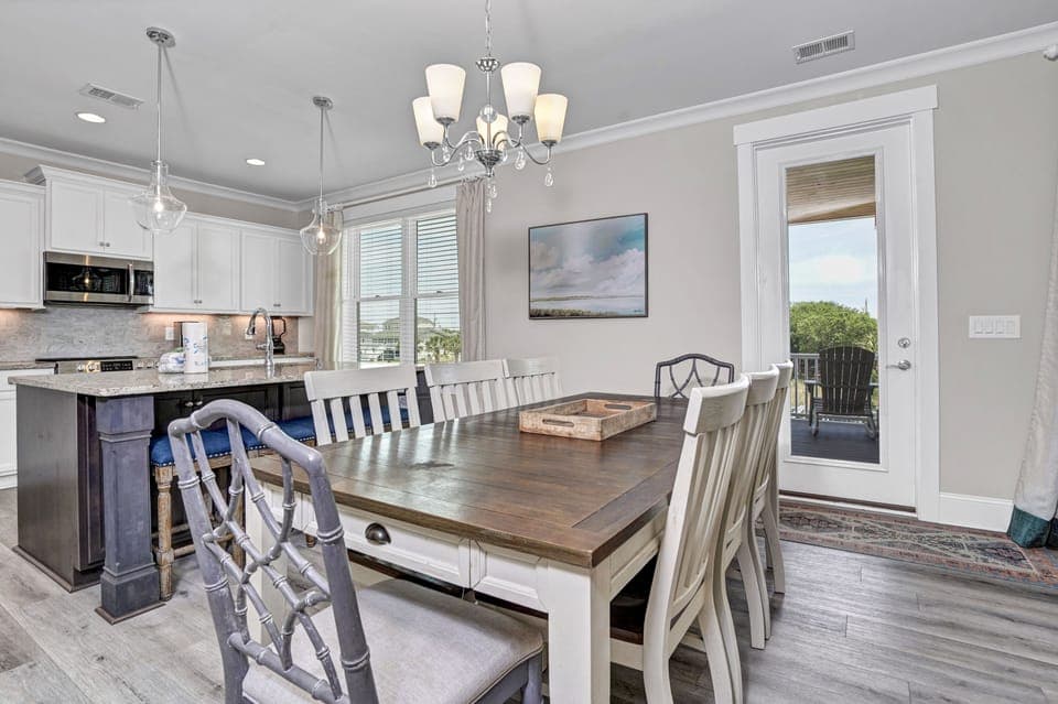 Your elegant dining space features a rustic wooden table beneath a stunning chandelier, with kitchen island seating and balcony doors flooding the room with natural light.
