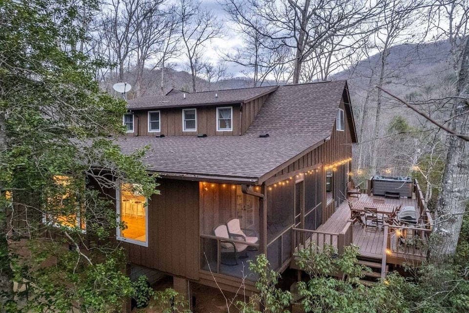 Multi-level exterior of Deer Creek Lodge featuring a private hot tub, outdoor dining area, and scenic wooded backdrop.