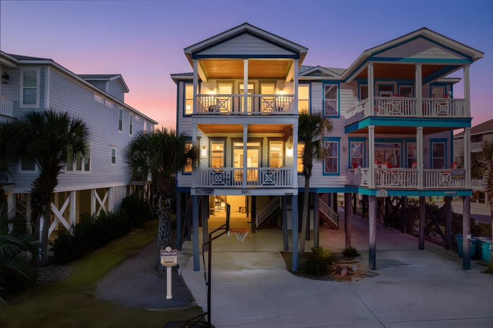 Beach house neighborhood with colorful coastal homes elevated on stilts, featuring covered parking and sunset sky backdrop.