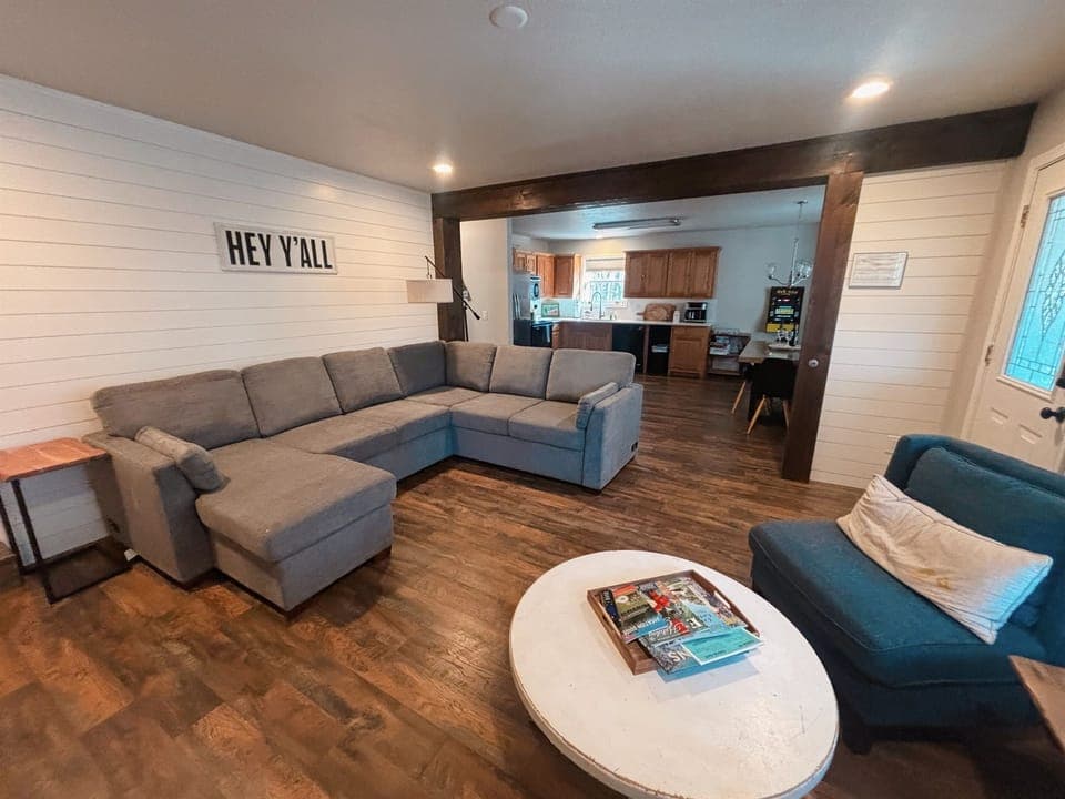 Sprawl out and unwind in this bright, open living space featuring a plush sectional, warm wood floors, and seamless flow into the kitchen—perfect for movie nights, morning coffee, and making unforgettable memories together.