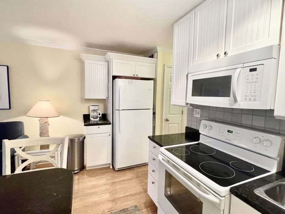 Compact kitchen with white cabinets, black countertops, refrigerator, stove, microwave, and coffee maker. A small dining area is visible with a wooden chair and lamp.