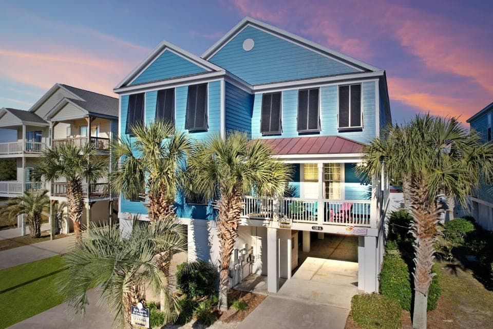 Charming coastal rental with vibrant blue siding, surrounded by swaying palm trees under a stunning sunset sky.